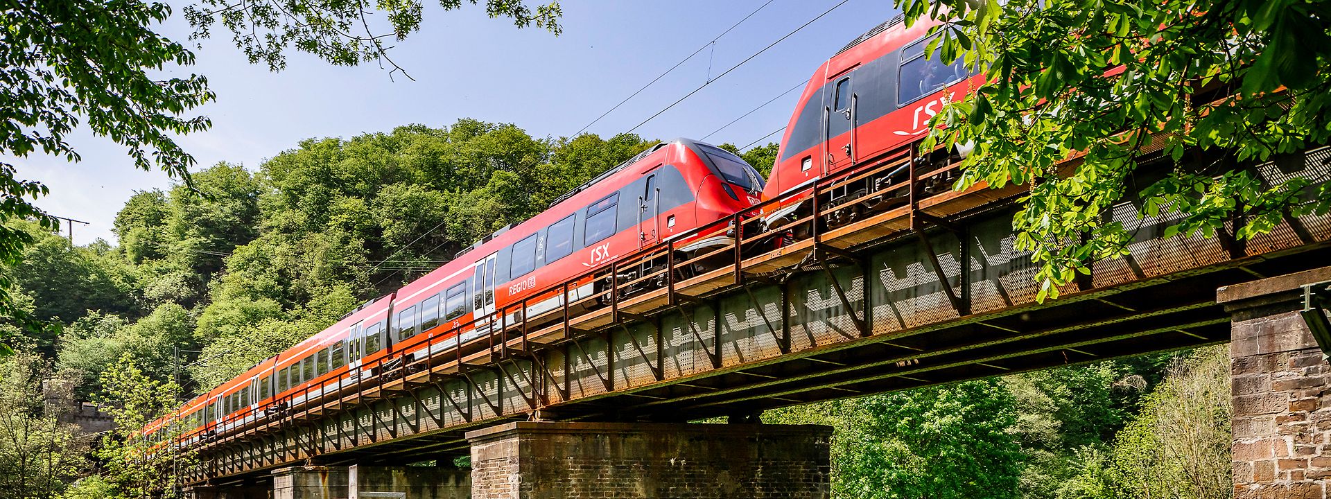 Quelle: DB AG / Smilla Dankert Der RSX fährt auf einer Gleisbrücke bei blauem Himmel durch eine grüne Naturlandschaft.