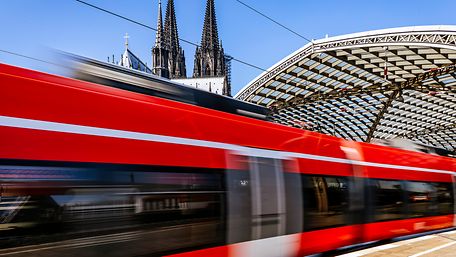 Ein roter Zug des RSX fährt in den Kölner Hauptbahnhof ein, im Hintergrund ist das Dach des Kölner Doms erkennbar. 