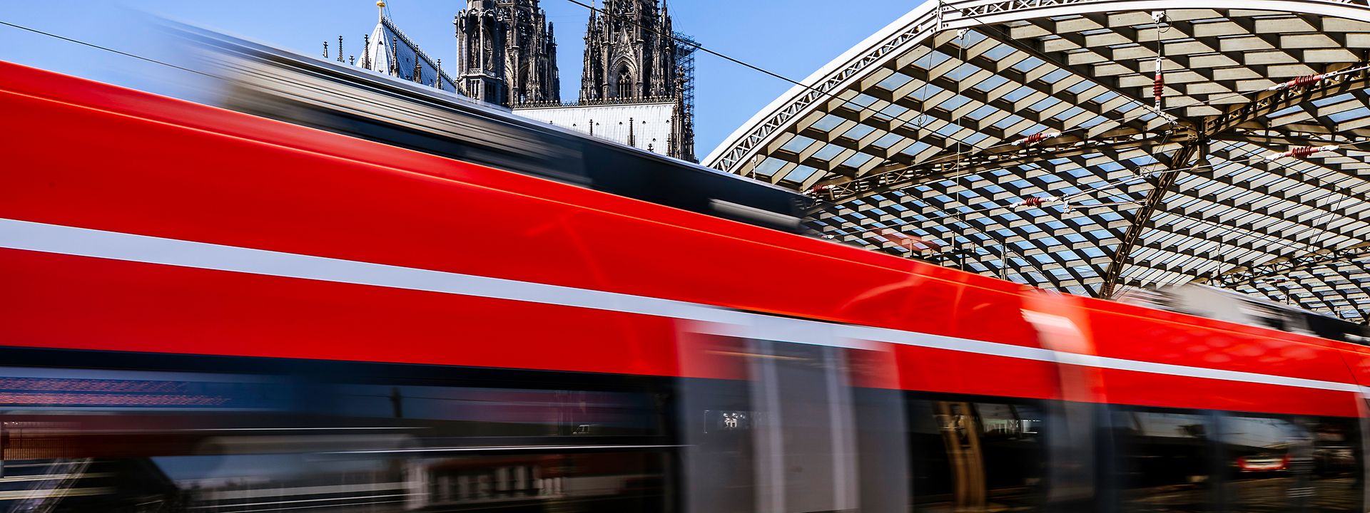 Ein roter Zug des RSX fährt in den Kölner Hauptbahnhof ein, im Hintergrund ist das Dach des Kölner Doms erkennbar. 