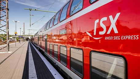 Ein roter Zug des RSX steht an einem Gleis unter freiem Himmel, das Logo des RSX ist am Wagen gut erkennbar