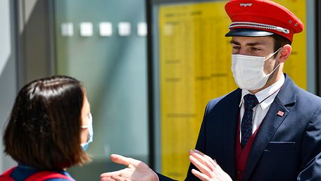 Ein junger Servicemitarbeiter der Deutschen Bahn mit Mund-Nasen-Schutz im Gespräch mit einer Reisenden