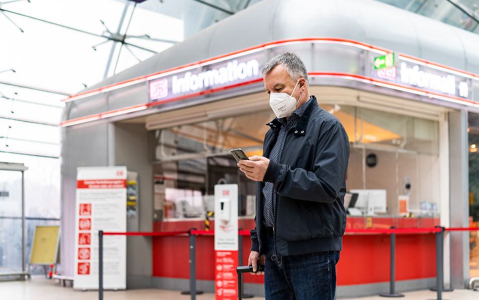 Ein Reisender informiert sich vor einer DB Information per Smartphone zu den Bahnfahrten.