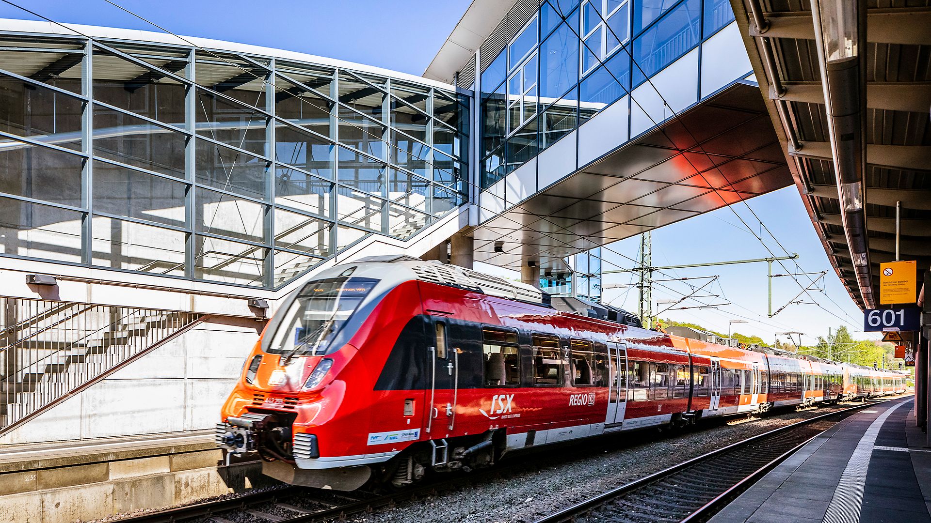 Entlangfahrender RSX-Zug in einer modernen Bahnhofsumgebung, welcher unter einer Übergangsbrücke hindurchfährt.